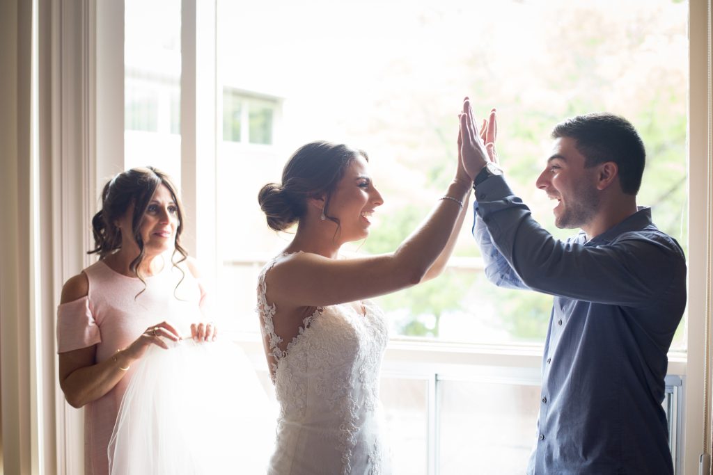 Bride and brother of the bride, giving each other high fives on her wedding day while the mother of the bride watches