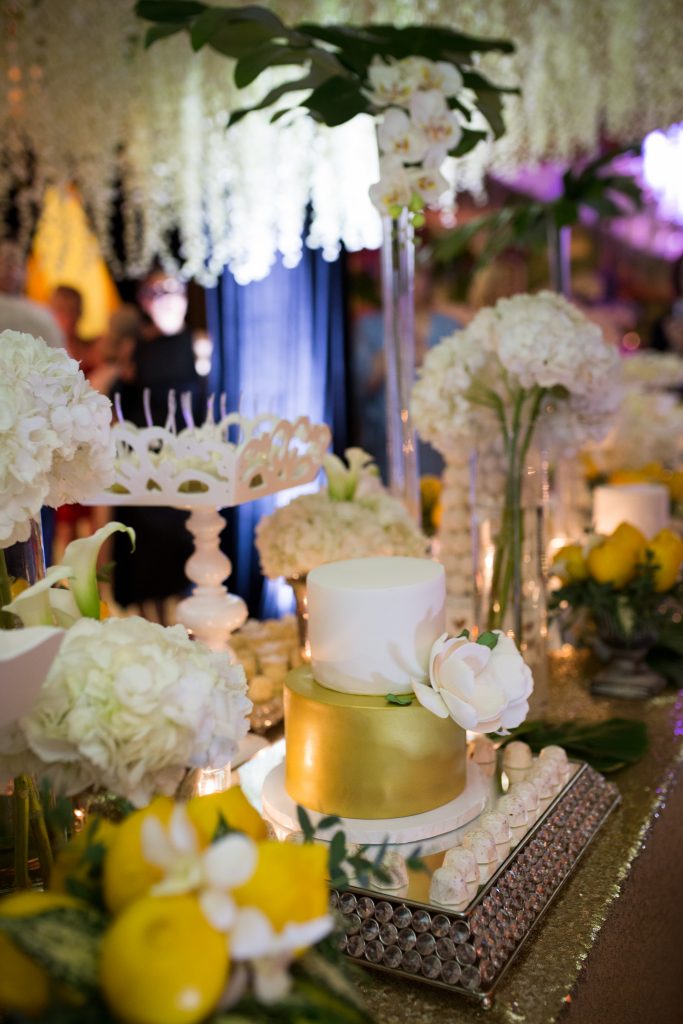 sweet table with mini wedding cake sourrounded by white flowers greenery and lemons with gold table cloth